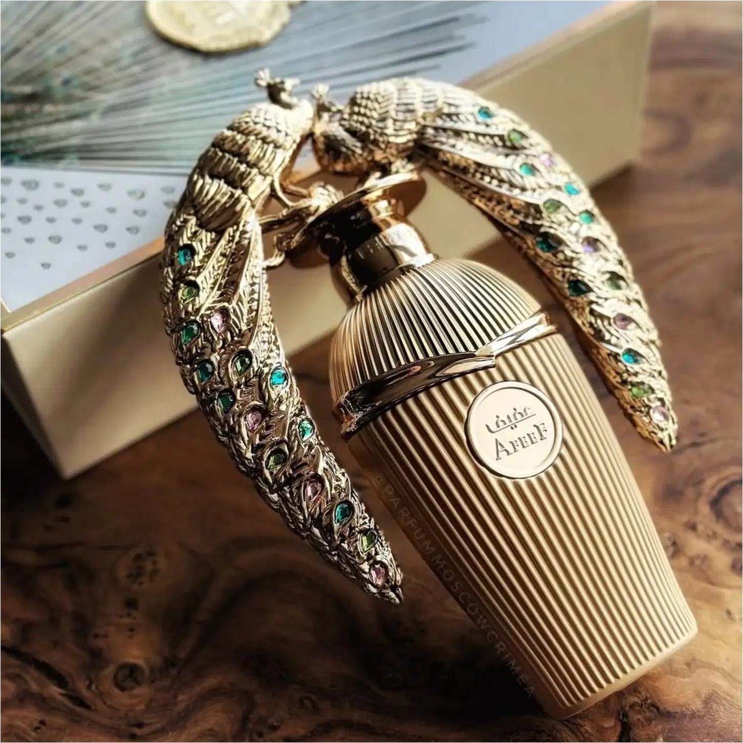 Frasco de perfume Lattafa Affee com embalagem decorativa em metal dourado, adornada com pedras coloridas em formato de penas. O rótulo do produto é visível na parte frontal do frasco.