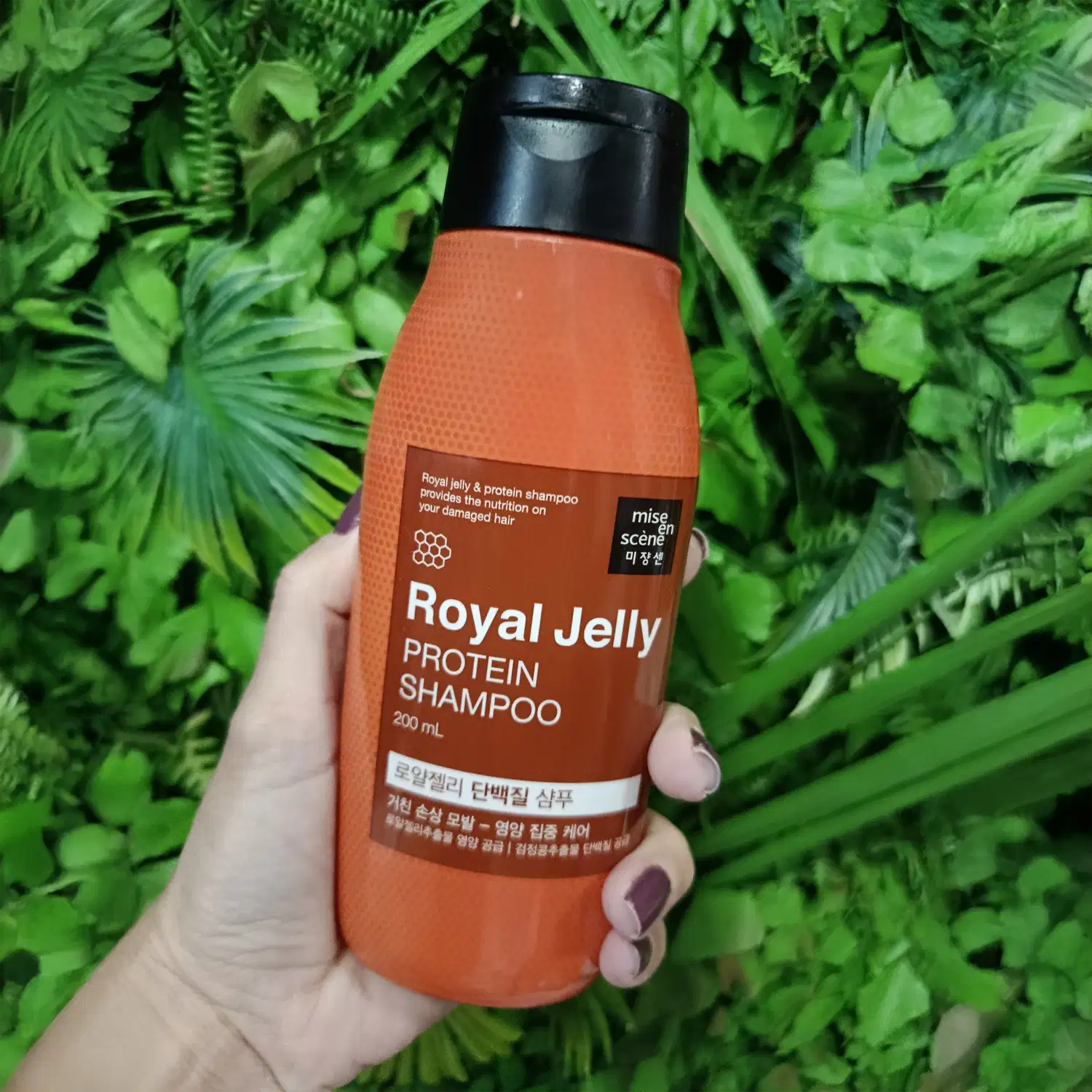 Frasco de shampoo de geléia real e proteína da marca Mise en Scène. O recipiente possui uma superfície laranja brilhante, com rótulo em preto e marrom destacando os ingredientes principais.