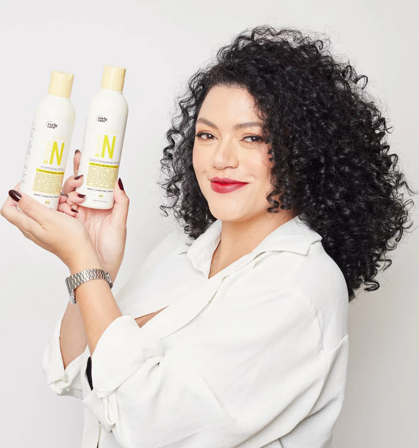 A imagem mostra dois frascos de produtos para cabelos cacheados da marca Curly Care. Os frascos são brancos e alongados, com tampas amarelas. Os frascos estão sendo segurados por uma mulher de cabelos cacheados escuros e volumosos.