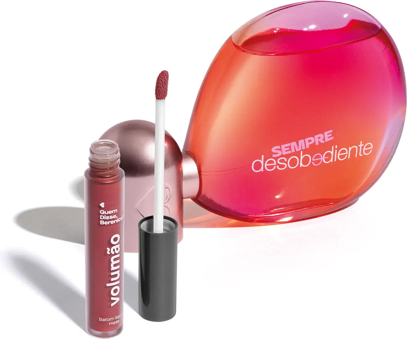 Imagem do Eau de Parfum Sempre Desobediente com vidro transparente em formato oval, líquido rosa e letras brancas e batom líquido Volumão Roseli na cor rosa em embalagem transparente e aplicador macio também marrom. O fundo é branco com sombreado.