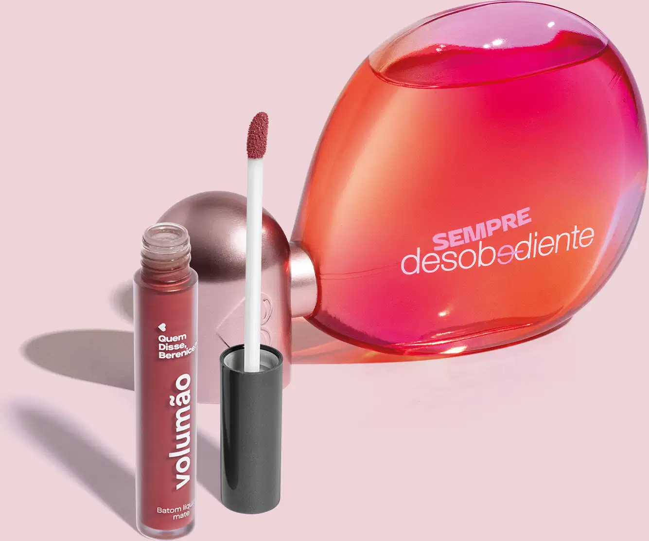 Imagem do Eau de Parfum Sempre Desobediente com vidro transparente em formato oval, líquido rosa e letras brancas e batom líquido Volumão Roseli na cor rosa em embalagem transparente e aplicador macio também marrom. O fundo é bege com sombreado.