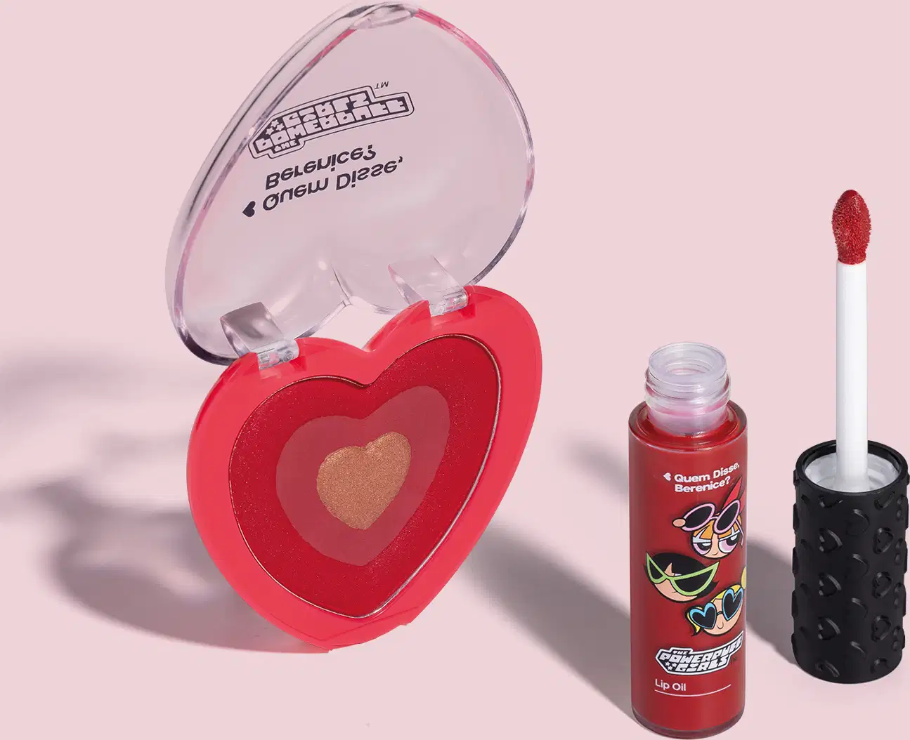 Imagem de dois itens da marca QDB As Meninas Super Poderosas, sendo um blush compacto rosa em formato de coração e um lip oil vermelho com aplicador em forma de esponja na cor vermelha. O fundo da imagem é neutro com sombreado.