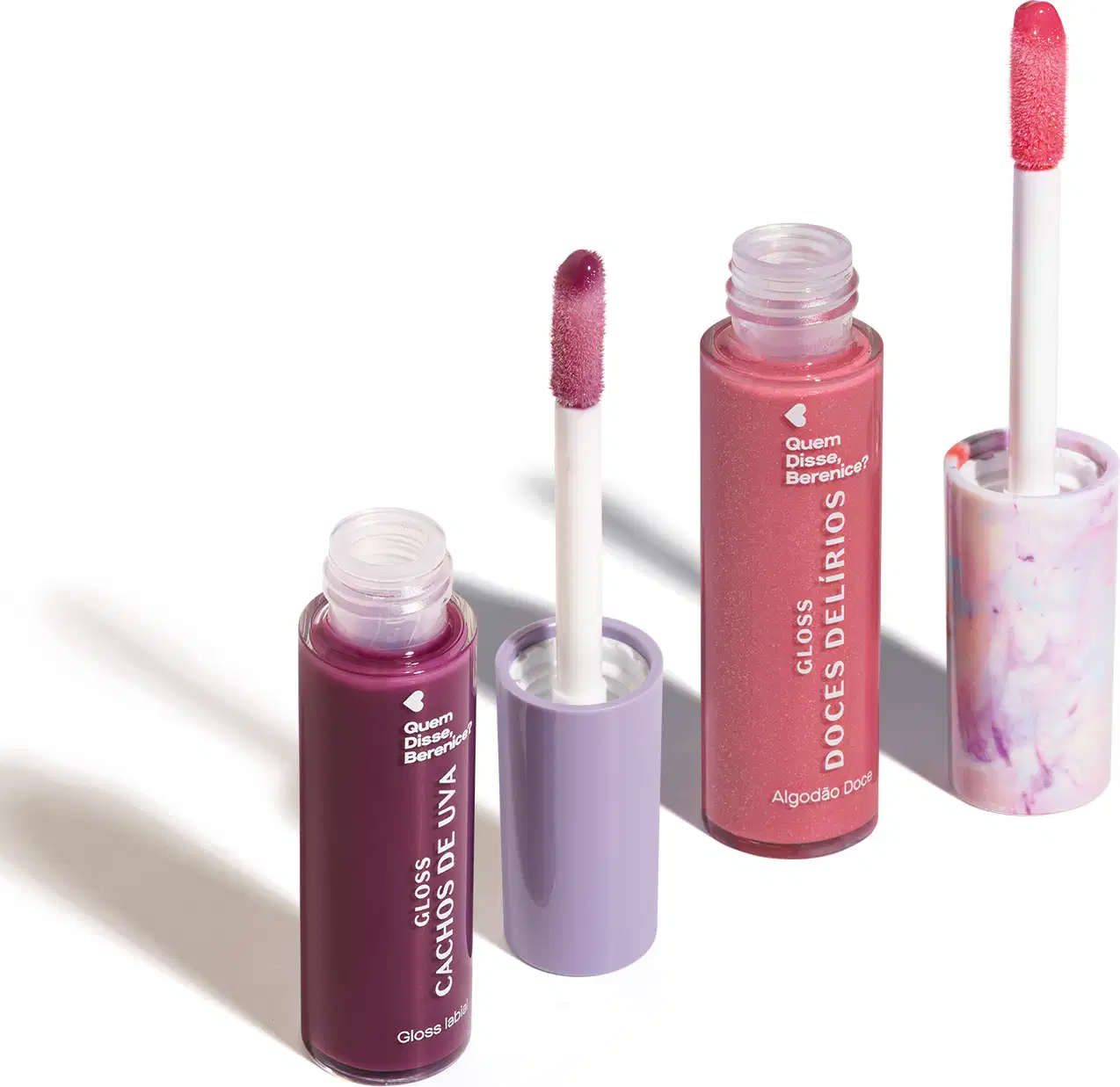 Imagem de dois frascos de gloss labial, um roxo com título Cachos de Uva e outro rosa com título Doces Delírios. Ambos em recipiente transparente com aplicador em forma de esponja na mesma cor do gloss. O fundo da imagem é branco com sombreado.