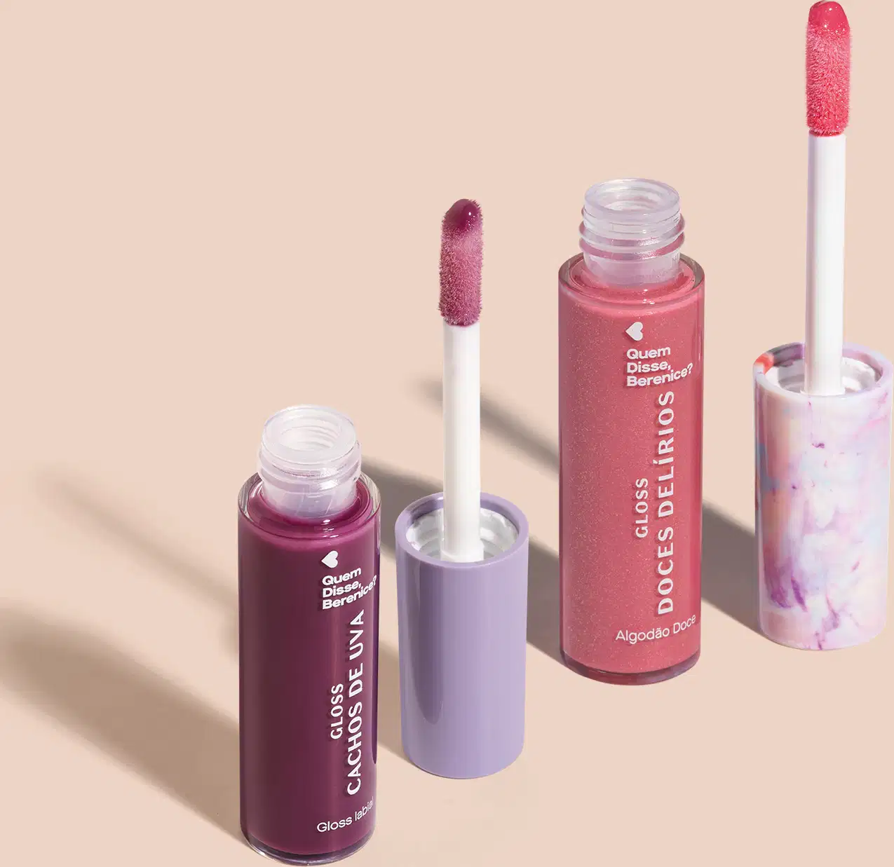 Imagem de dois frascos de gloss labial, um roxo com título Cachos de Uva e outro rosa com título Doces Delírios. Ambos em recipiente transparente com aplicador em forma de esponja na mesma cor do gloss. O fundo da imagem é neutro com sombreado.