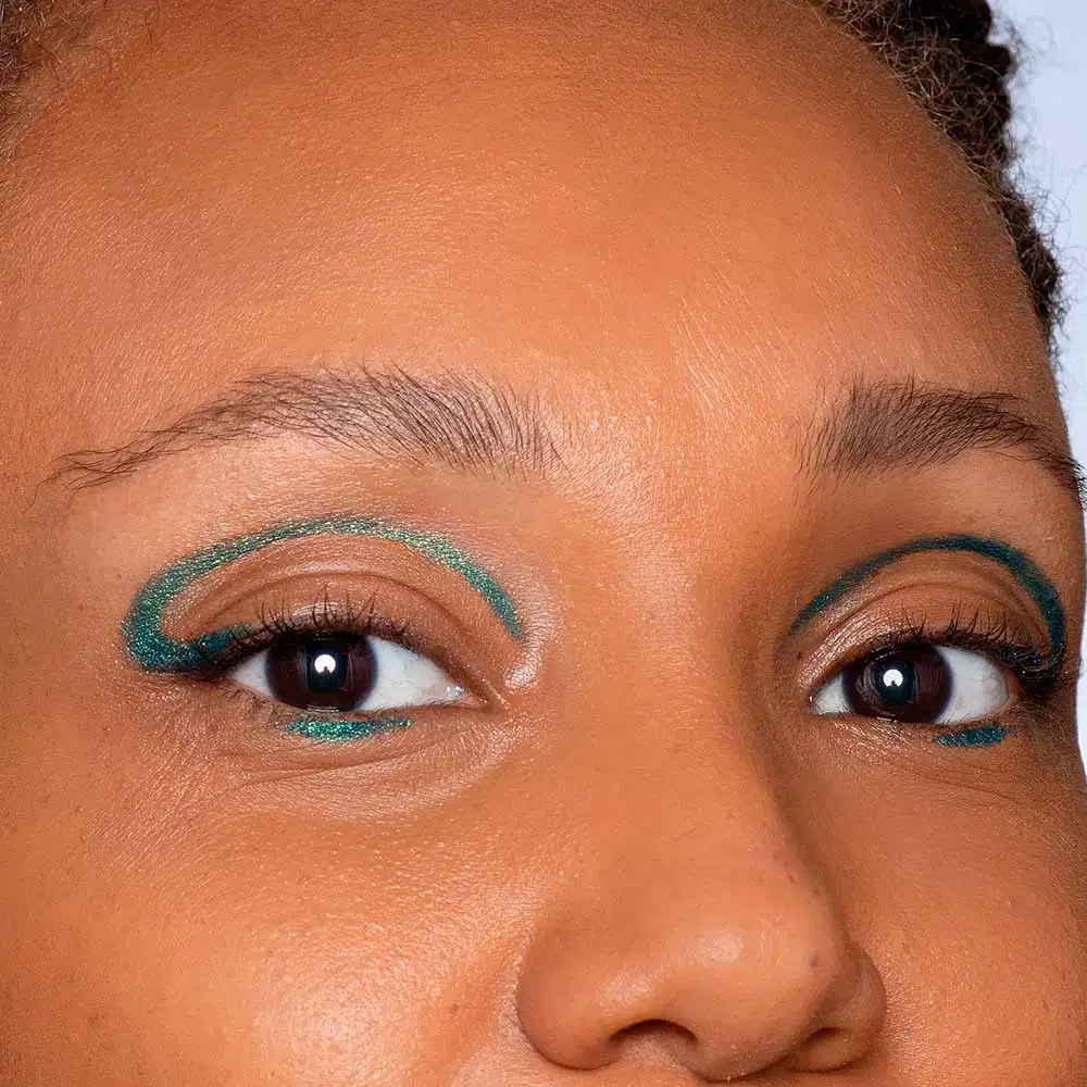 Olhos de uma pessoa com maquiagem colorida em tons de marrom e verde. A imagem mostra os olhos de perto, com sobrancelhas bem definidas e cílios destacados. A maquiagem parece ser aplicada com cuidado e detalhe, criando um visual elegante e expressivo.