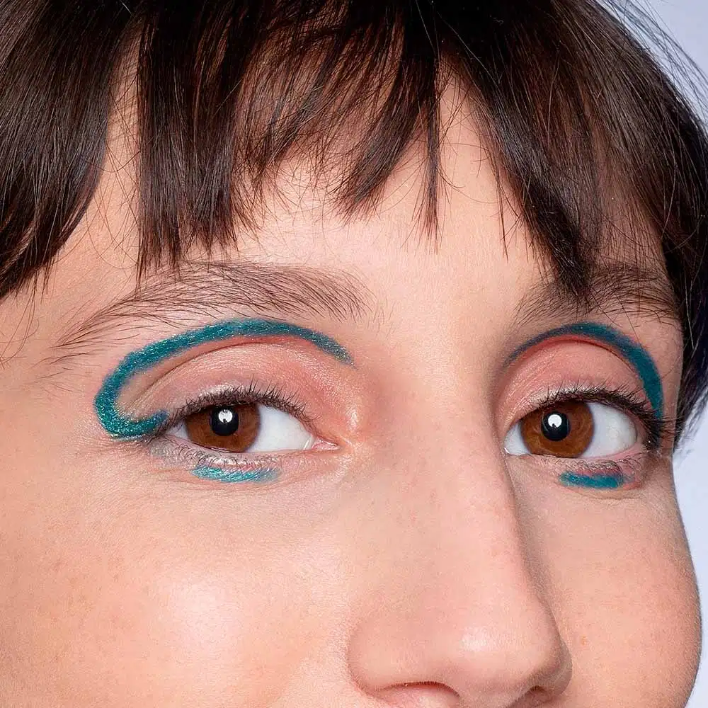 Olhos castanhos de uma pessoa com maquiagem colorida em tons de verde e marrom. A imagem mostra o rosto próximo, com foco nos olhos e sobrancelhas.