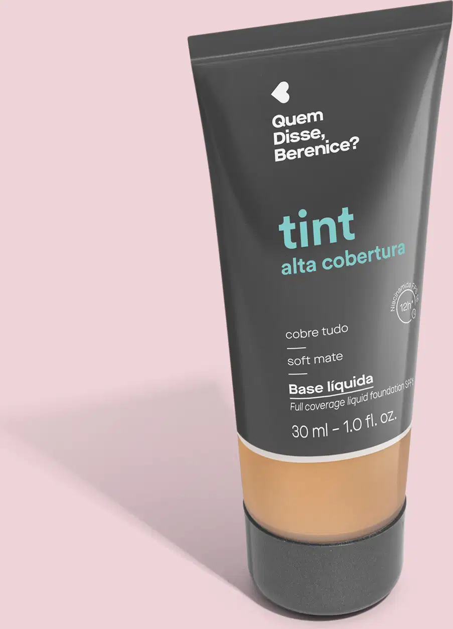 A imagem mostra a Base Tint Alta Cobertura da Quem Disse, Berenice? Embalagem cinza escura com parte inferior transparente para mostrar a cor do produto sobre fundo rosa.