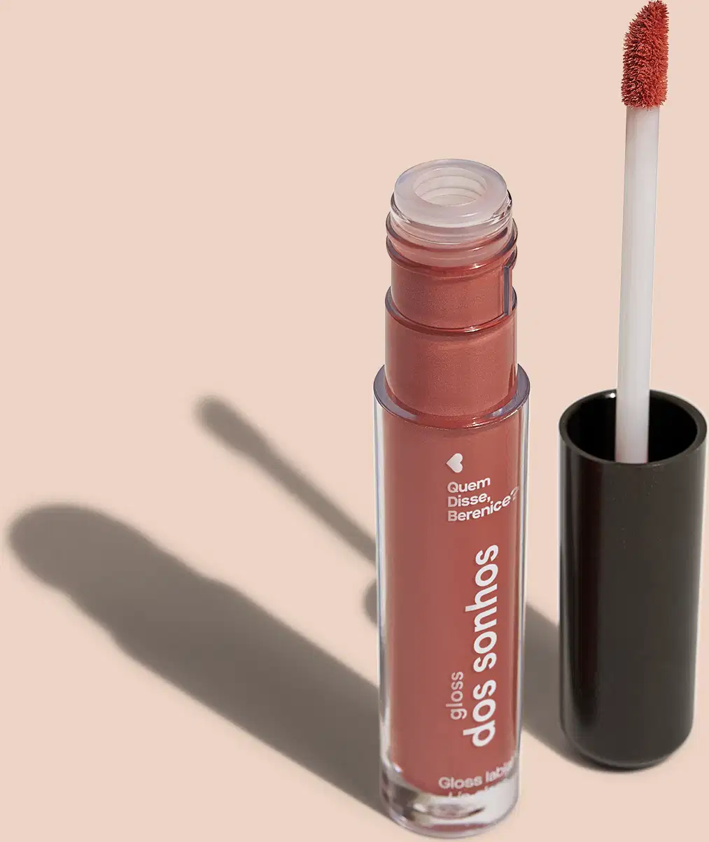 Frasco cilíndrico transparente contendo um gloss labial marrom. Rótulo na embalagem com o nome do produto "Gloss dos Sonhos" e a descrição "Quem disse, berence?". Ao lado, a tampa preta do produto, que acompanha um aplicador de espuma.