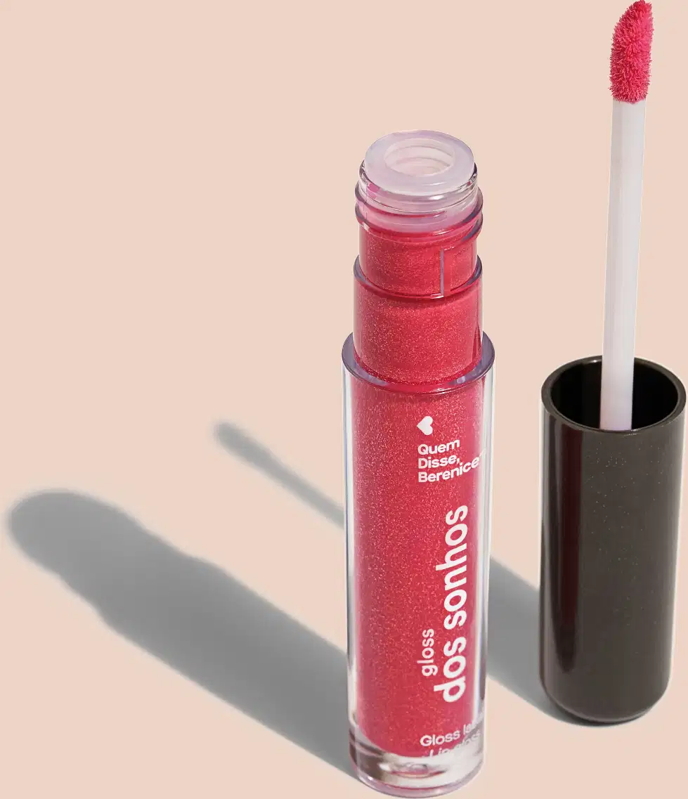 Frasco cilíndrico transparente contendo um gloss labial rosa brilhante Rótulo na embalagem com o nome do produto "Gloss dos Sonhos" e a descrição "Quem disse, berence?". Ao lado, a tampa preta do produto, que acompanha um aplicador de espuma rosa.