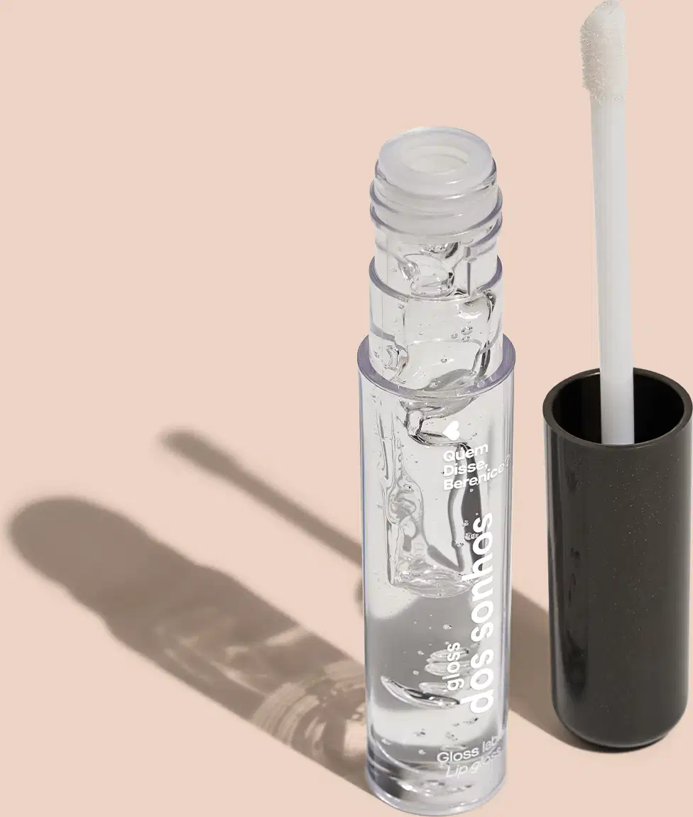 Gloss labial transparente em frasco cilíndrico de vidro com tampa plástica branca. Ao lado, um aplicador de maquiagem em formato de pincel com cabo preto. Embalagem com o texto "gloss dos sonhos" visível. Cores predominantes são transparente e preto.