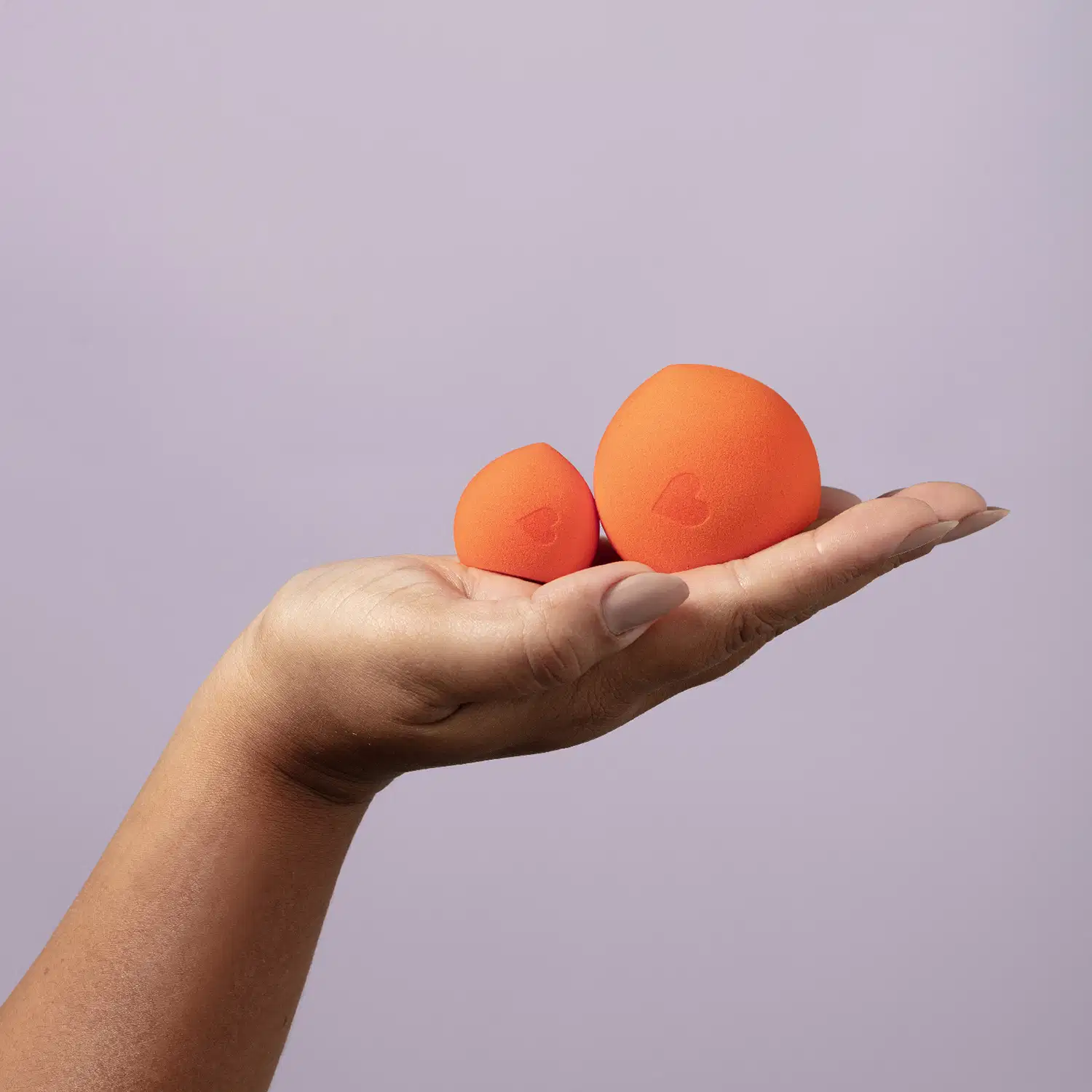 A imagem mostra duas esponjas de cor laranja, que parecem ser produtos de beleza da marca Quem Disse, Berenice?. Ambos estão sendo sendo segurados por uma mão, com um fundo liso de cor lilás.