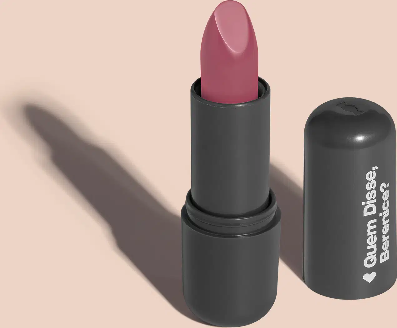 Batom de cor rosa claro em embalagem preta com logotipo da marca Quem Disse, Berenice? em destaque. O batom está aberto sobre superfície rosa com iluminação que cria uma longa sombra projetada atrás da embalagem.