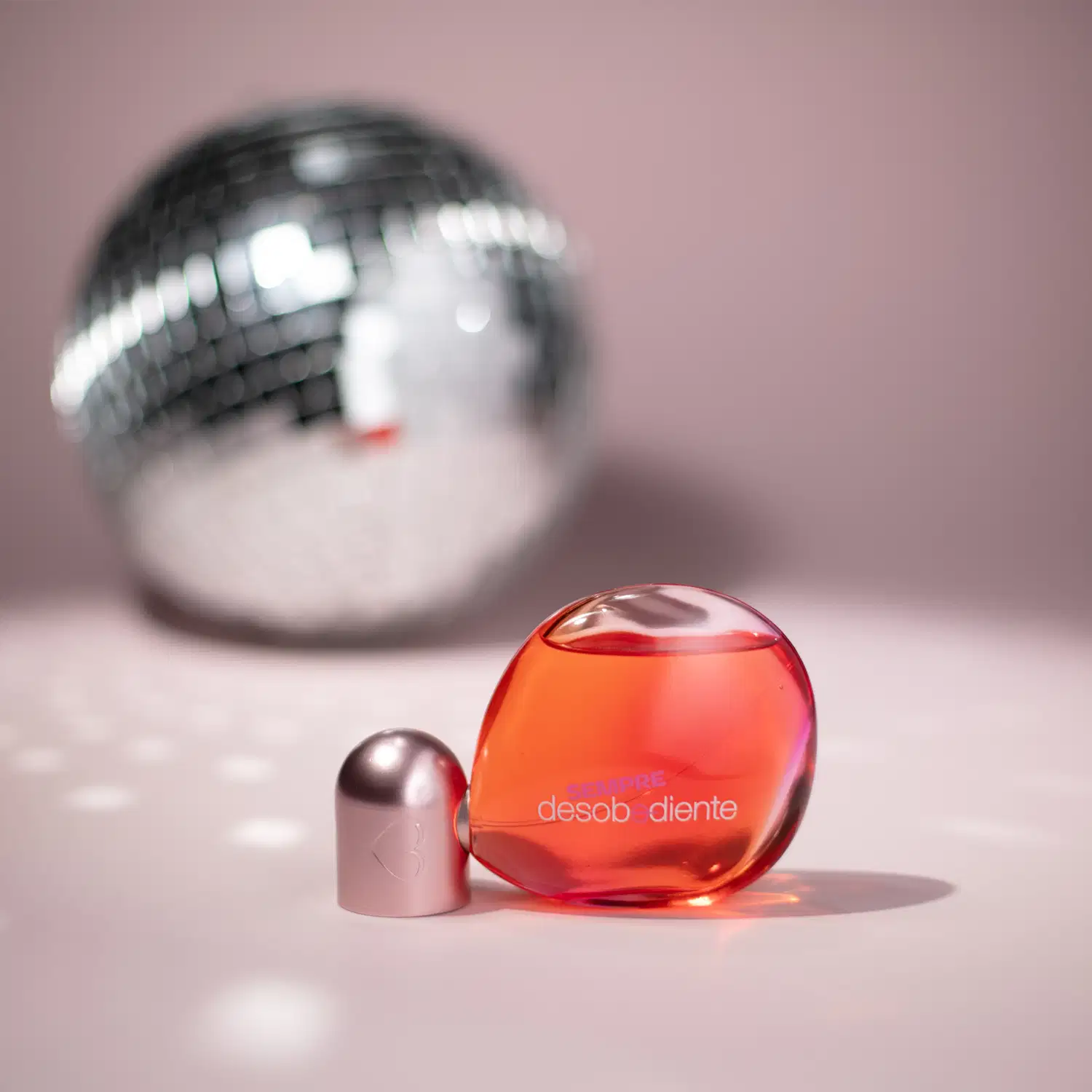 Frasco de Eau de Parfum Desobediente vermelho e rosa com texto em destaque, sobre superfície rosa, com bola prateada ao fundo.