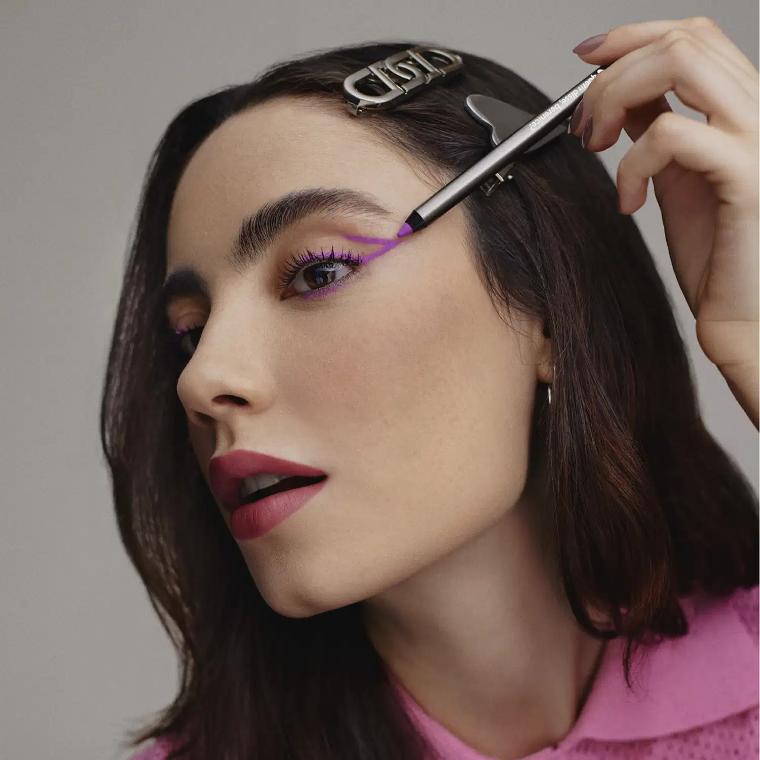 A imagem mostra uma modelo usando maquiagem de olhos em tons de roxo vibrante. Ela tem sobrancelhas bem delineadas e batom vermelho escuro nos lábios. Seu cabelo castanho escuro está penteado para trás, permitindo ver claramente seu rosto.
