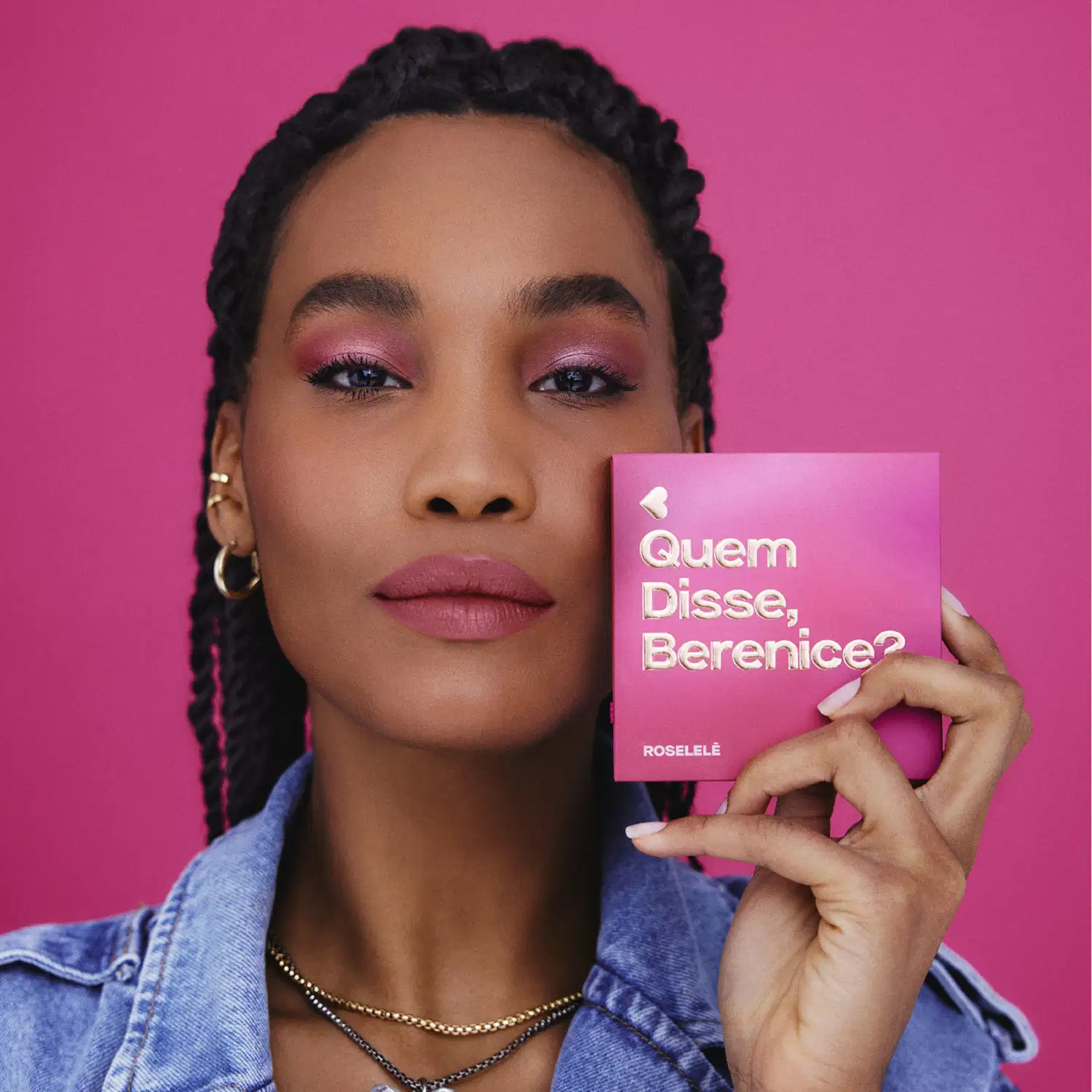 A paleta de maquiagem é segurada por uma pessoa com penteado de tranças, maquiagem e acessórios, em frente a um fundo rosa vibrante.
