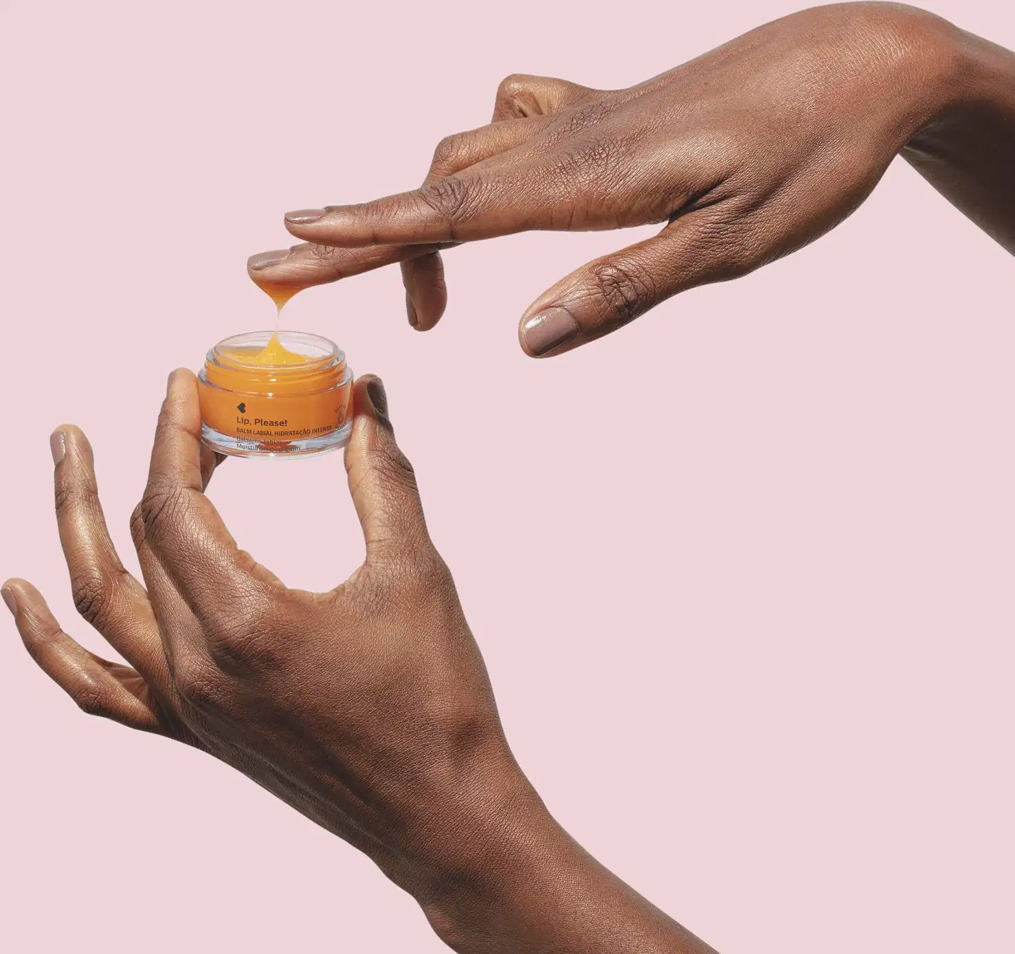 Mãos femininas, uma segurando um frasco aberto de balm labial de hidratação intensa Lip, Please! Quem Disse, Berenice? e a outra com uma pequena quantidade do produto em um dos dedos em fundo lilás claro.
