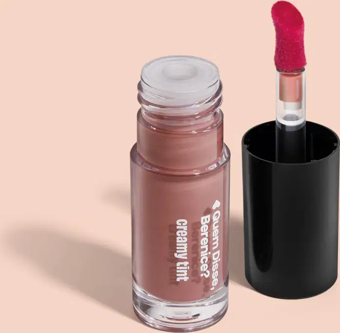 A imagem mostra um recipiente transparente de gloss labial da marca Quem Disse, Berenice? em tom de bege rosado, com aplicador vermelho, haste transparente, tampa preta, sobre superfície clara e neutra.