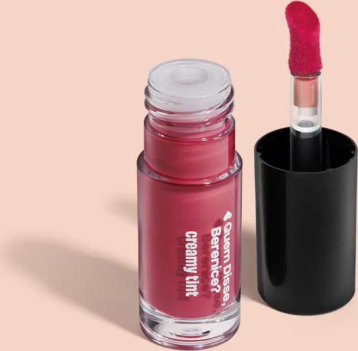 A imagem mostra um recipiente do gloss labial Quem Disse Berenice? em um tom rosado. A embalagem é transparente com aplicador vermelho, haste transparente e tampa preta, sobre fundo bege claro com leve sombreado.