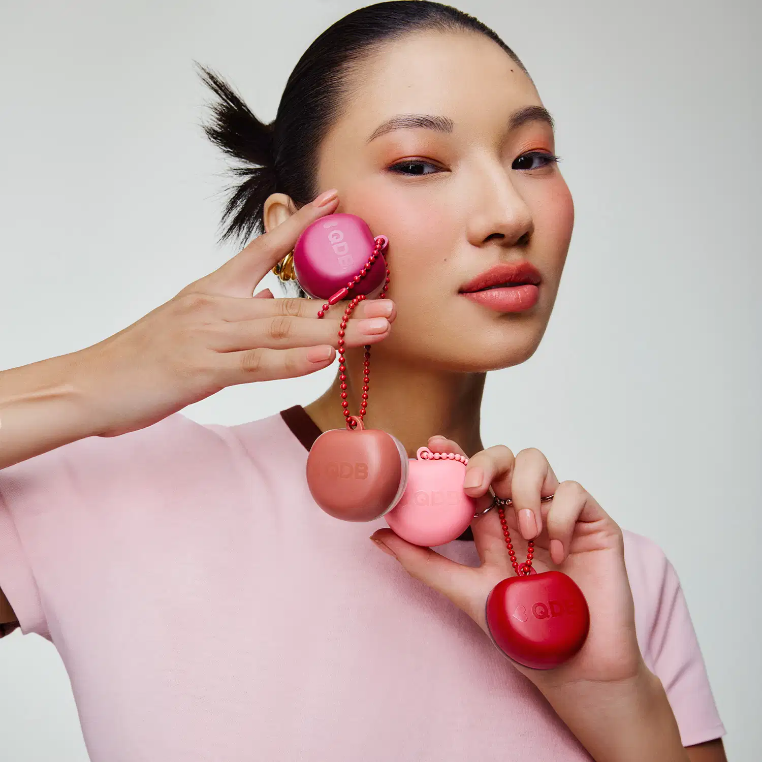 Uma pessoa segura quatro itens de perfumaria rosa com detalhes em vermelho. Os itens parecem ser pequenos frascos ou recipientes com cordões vermelhos. O fundo é branco, criando um ambiente minimalista e focado nos produtos.