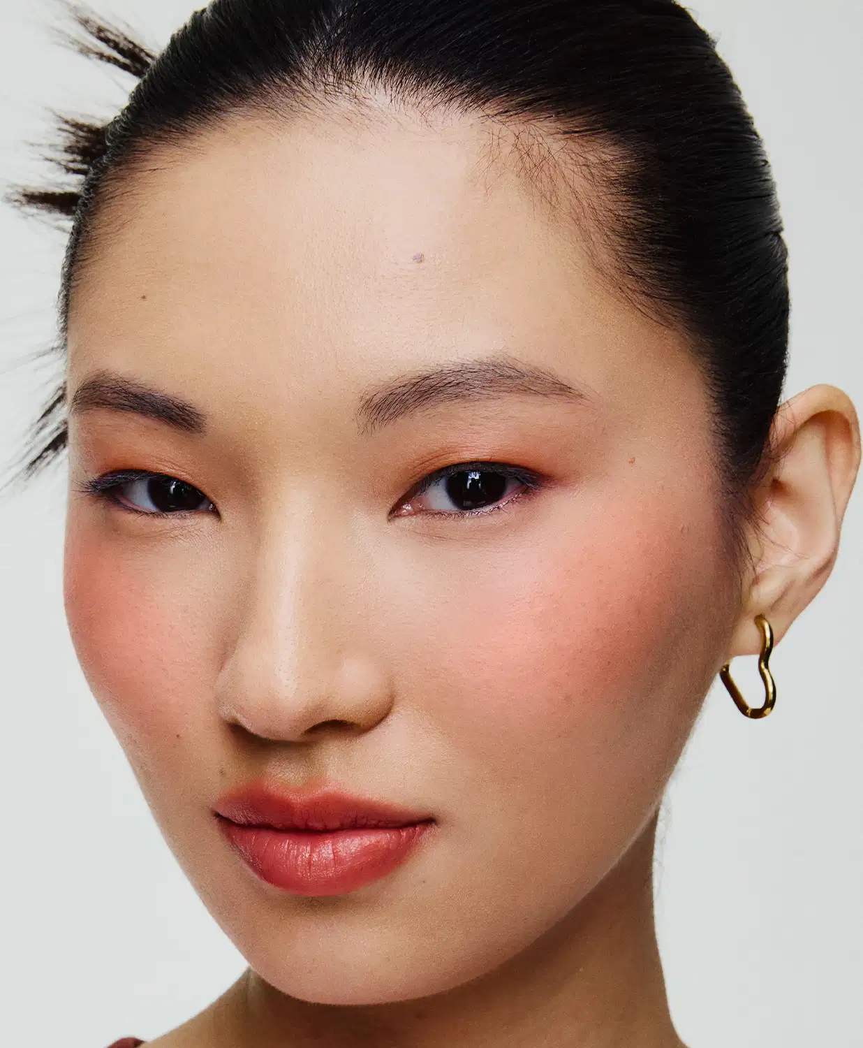 Rosto de uma mulher asiática com cabelo escuro e penteado elegante. Ela usa brincos dourados e maquiagem com tons de laranja e vermelho nos olhos e lábios. O fundo é branco, criando um ambiente minimalista e focado no rosto da modelo.