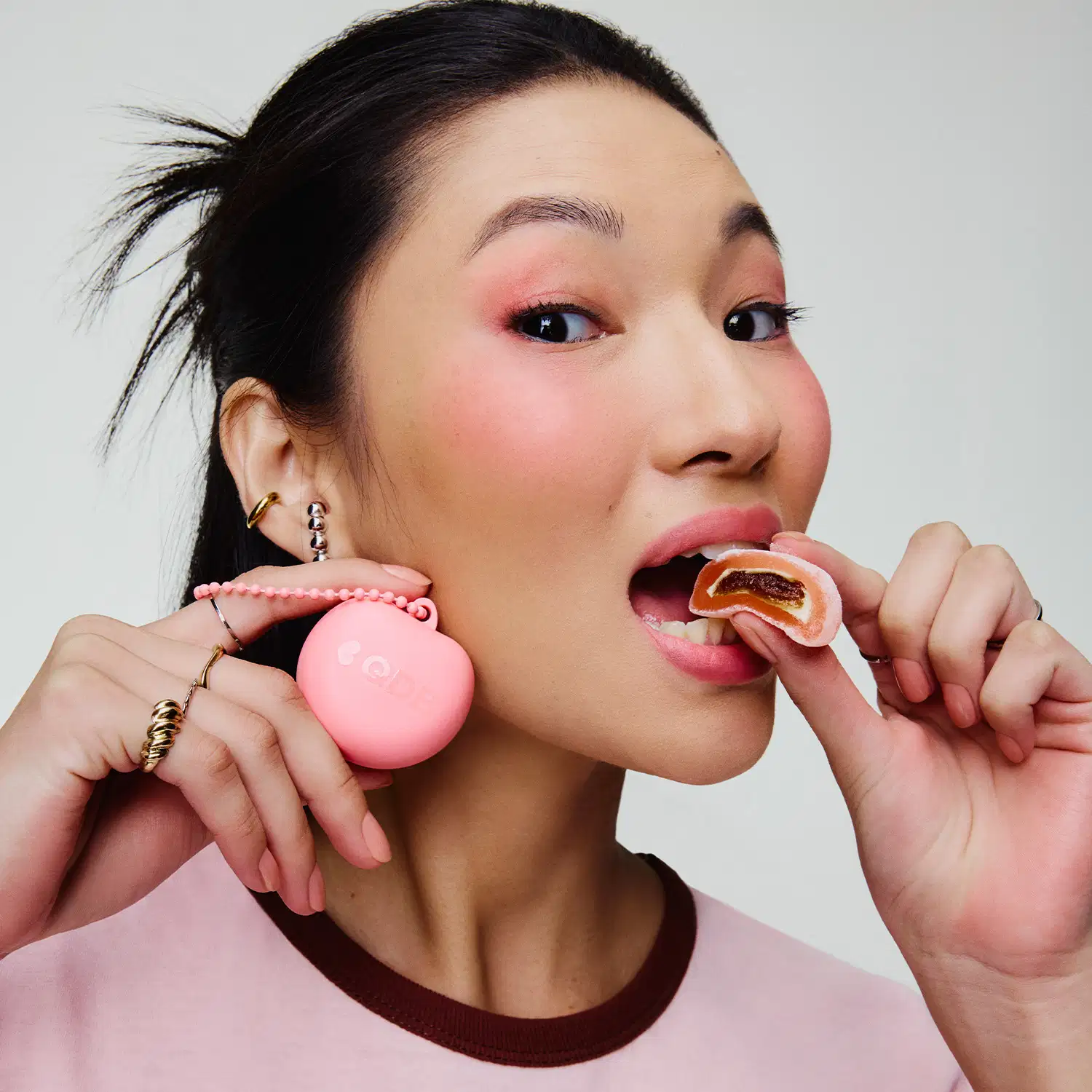 Uma mulher asiática segura um item de perfumaria rosa e um doce em suas mãos. Ela está usando joias douradas e sua expressão facial sugere que ela está apreciando o doce. O fundo é branco, criando um ambiente minimalista e focado no produto.