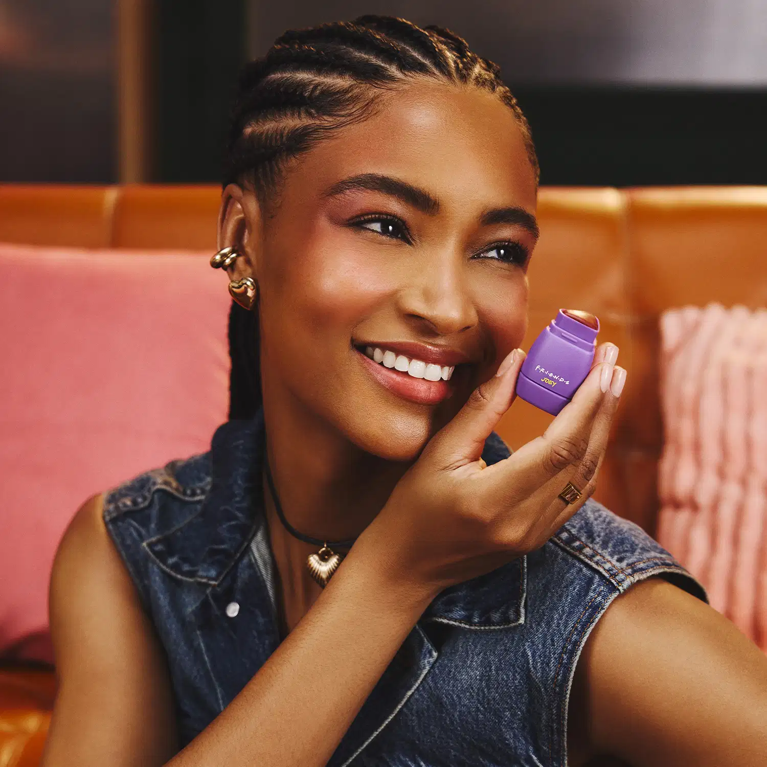 Uma mulher negra sorridente segurando um item de maquiagem roxo em um ambiente com decoração rosa e laranja. Ela usa tranças e acessórios dourados, vestindo uma jaqueta jeans.