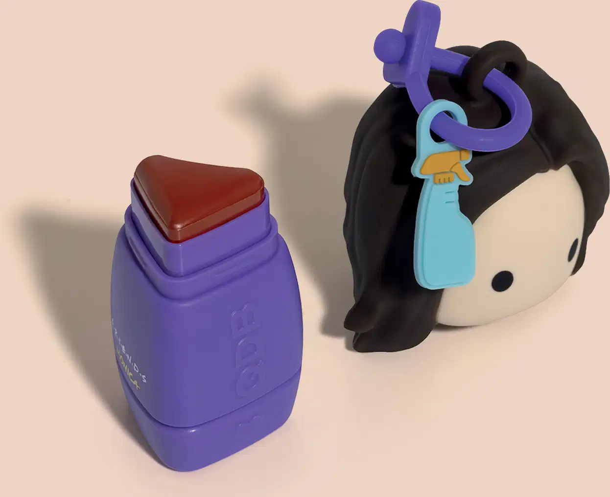 Batom de embalagem roxa em formato de coração. Ao lado, boneco preto com acessórios coloridos.