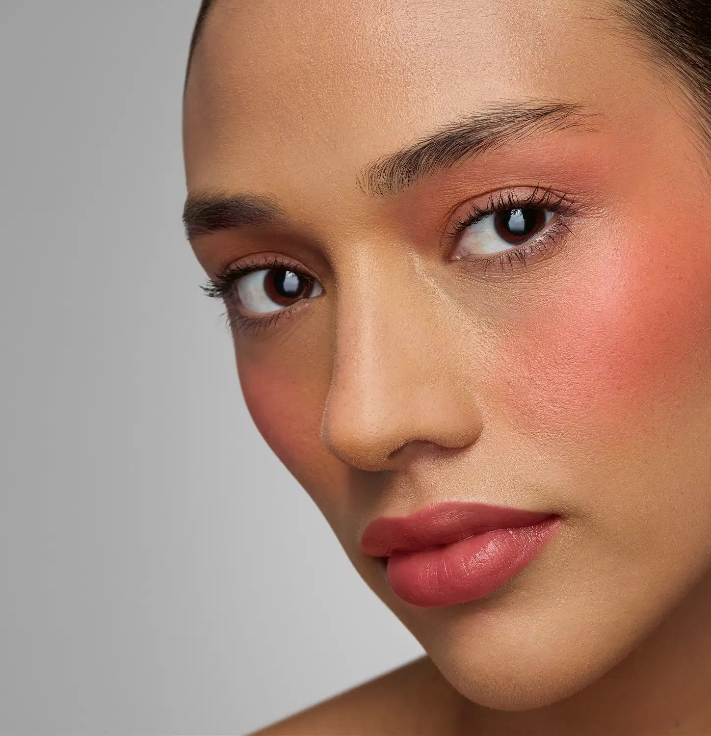 Rosto de uma pessoa com pele negra, olhos escuros e expressão séria. Lábios vermelhos e sobrancelhas bem definidas. Fundo cinza neutro que destaca os traços faciais. Imagem de alta qualidade que sugere um ambiente de beleza ou cosmética.