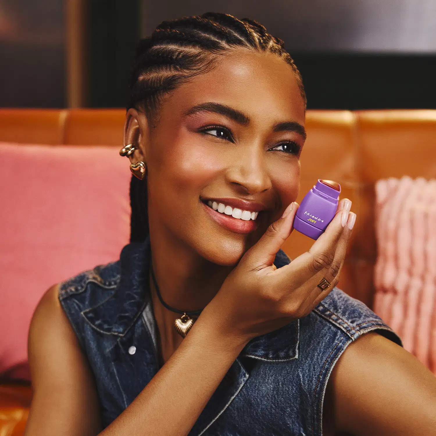 A pessoa sorri enquanto segura um item de maquiagem roxo em um ambiente com decoração rosa e laranja. Ela usa jaqueta jeans e acessórios dourados. O cenário sugere um ambiente informal e acolhedor, provavelmente relacionado a um produto de beleza.