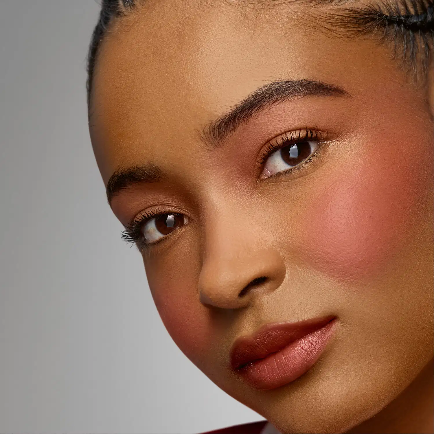 Rosto de uma mulher com pele negra, olhos escuros e expressão serena. Ela usa maquiagem sutil, com lábios vermelhos. O fundo é neutro, criando um ambiente de beleza e cuidado pessoal.