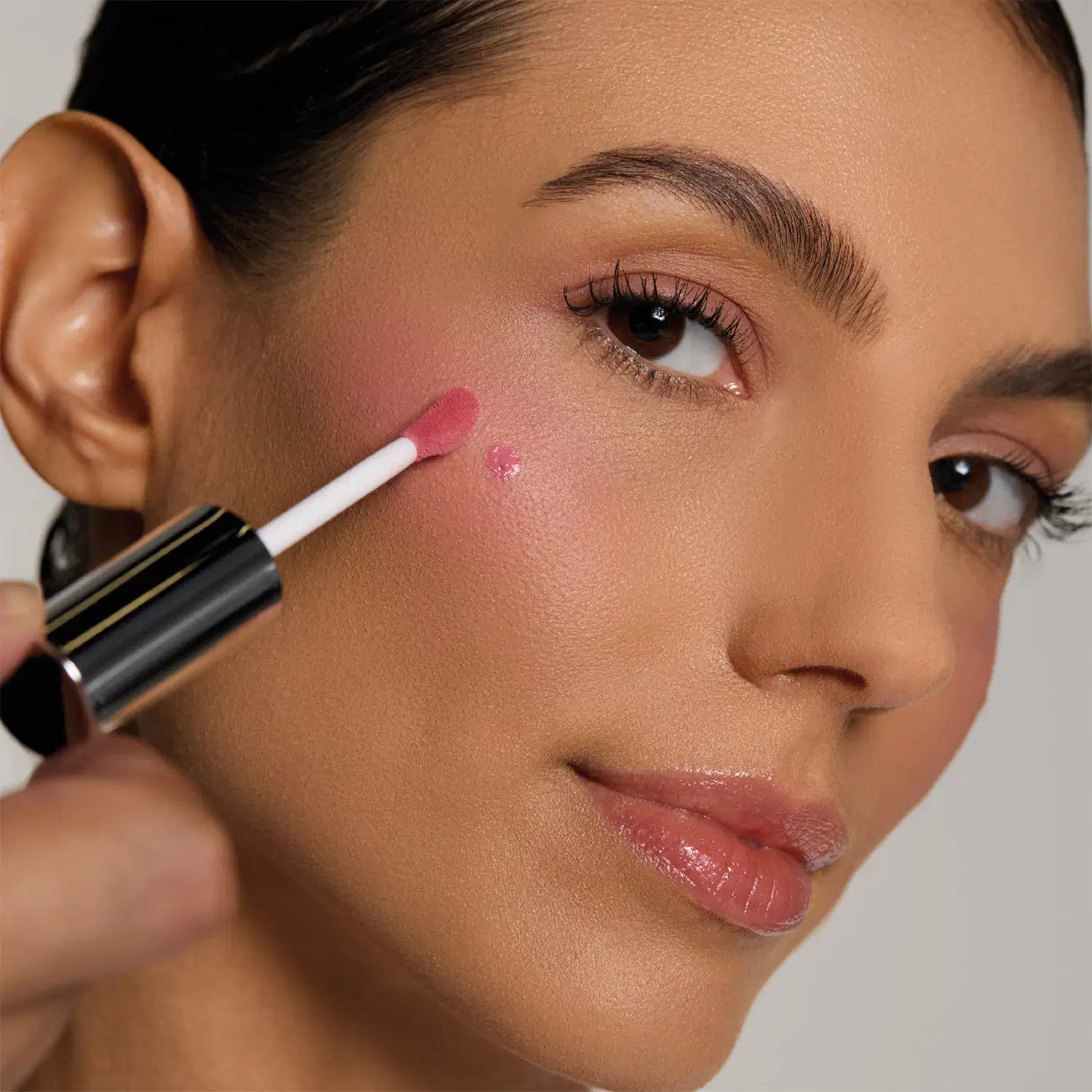 Uma mulher aplica um produto de maquiagem rosa em seu rosto, com fundo branco e iluminação suave.