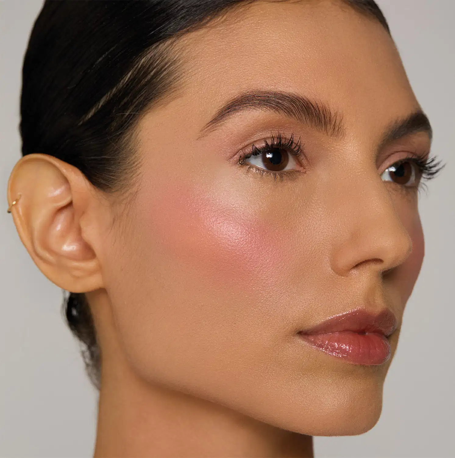 Rosto feminino com maquiagem natural, cabelos escuros e pele corada, em ambiente com fundo branco e iluminação suave.
