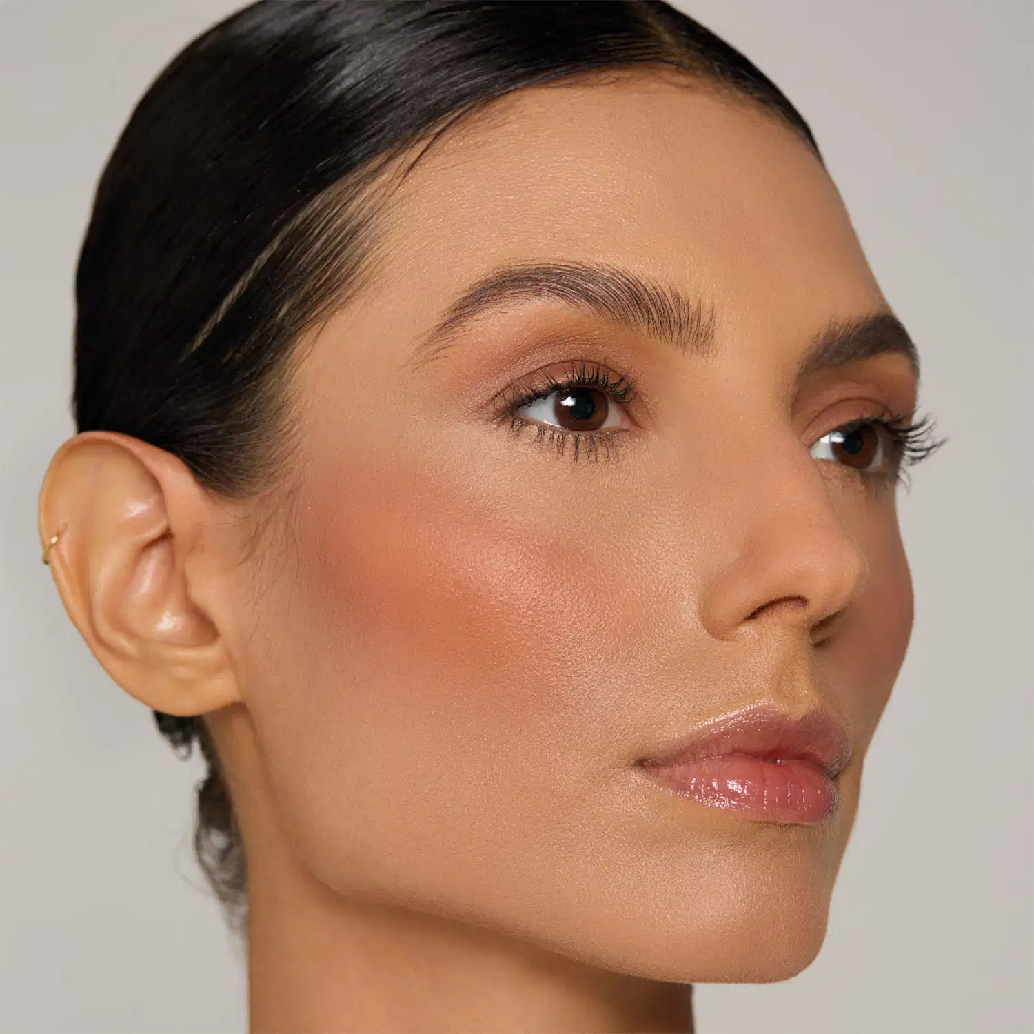 Rosto de mulher com maquiagem, cabelo escuro penteado para trás, fundo cinza claro, iluminação suave.