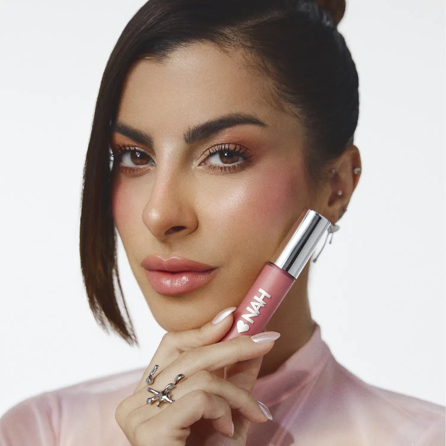 Mulher segurando um item de make rosa da marca Nah Cardoso em primeiro plano, com fundo branco e iluminação suave.