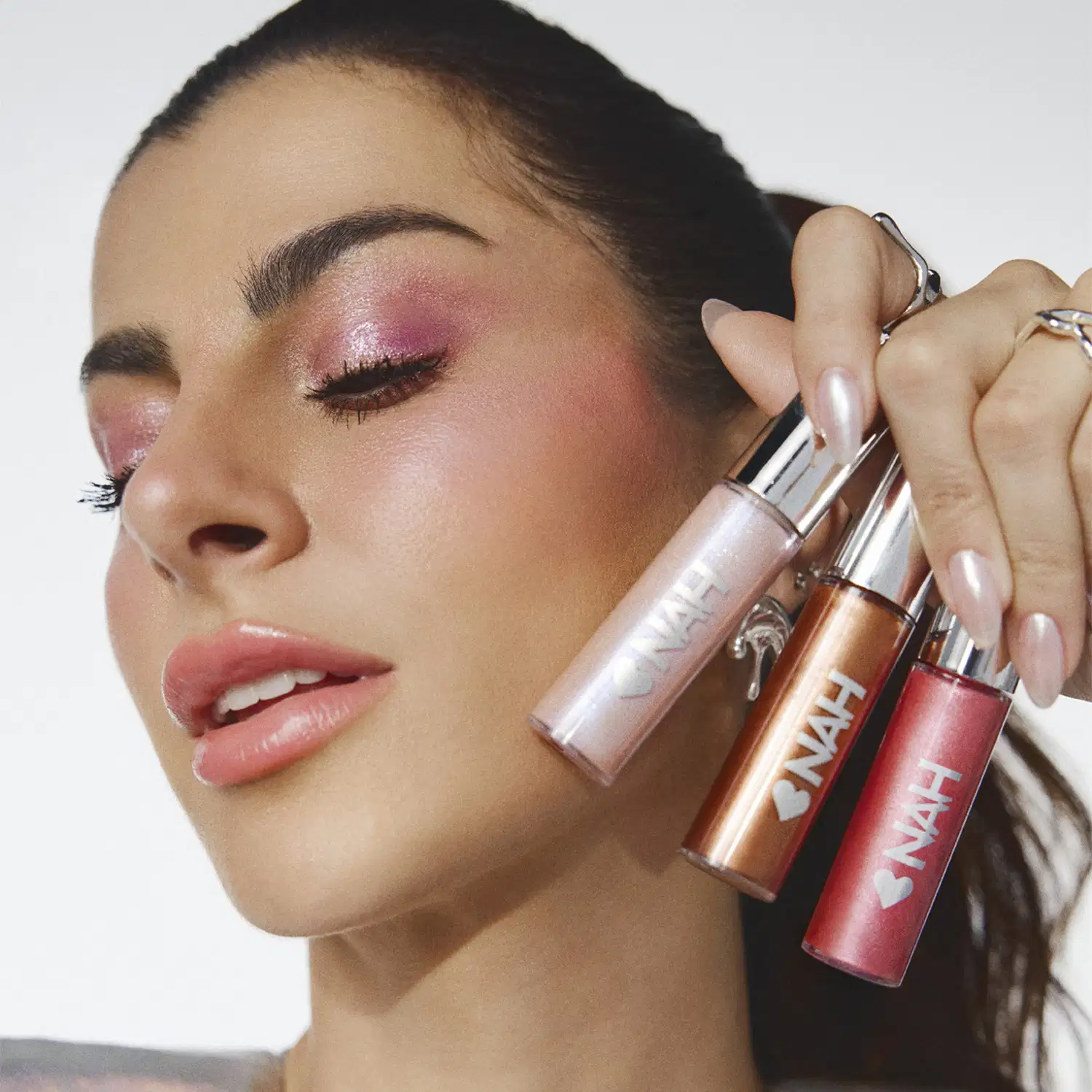 Três itens de make da marca Nah Cardoso em primeiro plano, com uma mulher segurando-os próximo ao rosto em um fundo branco.