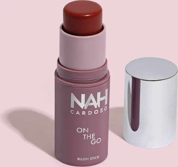 Produto de beleza em formato de bastão, com embalagem roxa e branca, exibindo o nome "NAH CARDOSO" e o texto "ON THE GO BLUSH STICK". O produto possui uma tampa prateada e uma cor marrom escuro.