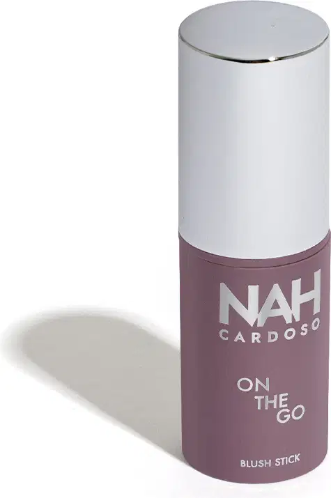 Produto de beleza em embalagem cilíndrica branca e roxa, com o nome "NAH CARDOSO" e a descrição "ON THE GO BLUSH STICK" impresso no rótulo. Trata-se de um blush em bastão, com design compacto e portátil, ideal para uso rápido e prático.