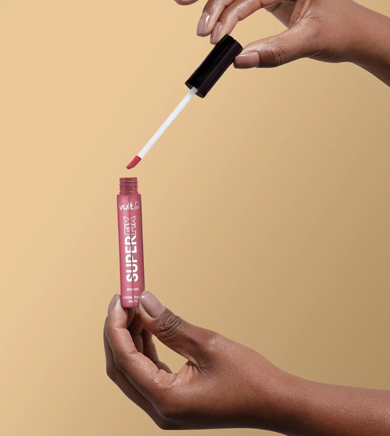 A imagem mostra uma mão segurando o produto aberto do Batom Líquido da marca Vult, na cor rosa queimado. A embalagem do produto é um tubo vermelho com o nome da marca "Vult" impresso.