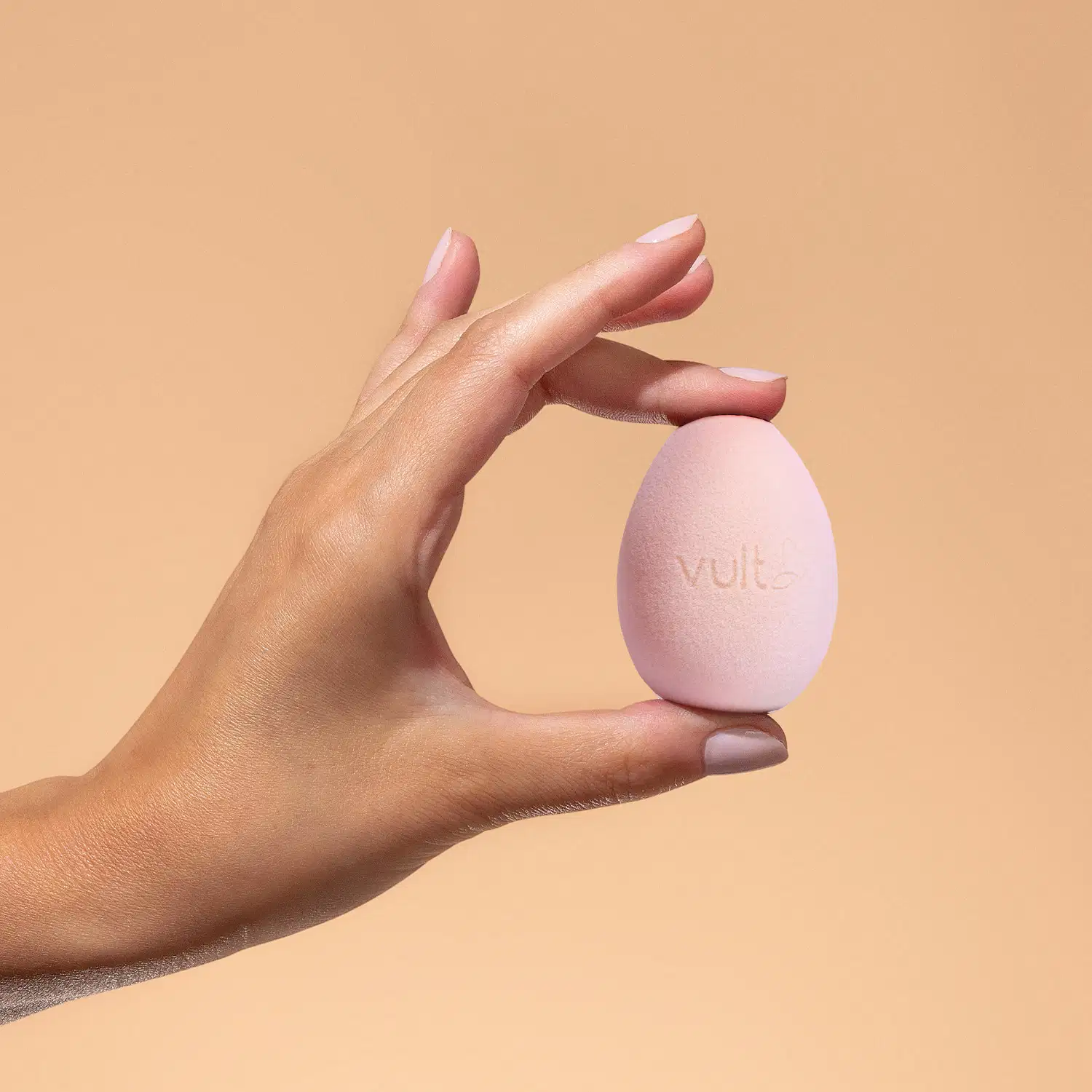 Mãos segurando uma esponja de maquiagem Vult em um fundo laranja, sugerindo um ambiente de cuidados com a pele e produtos de beleza.