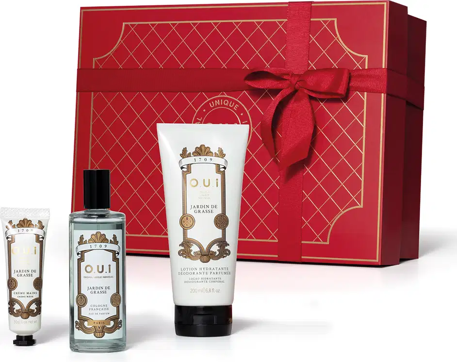 A imagem mostra um conjunto de produtos O.U.i em frente a uma bela caixa de presente vermelha. Ela traz um frasco com eau de parfum e dois tubos brancos com tampa preta, um menor com hidratante para as mãos e outro maior com hidratante para o corpo.