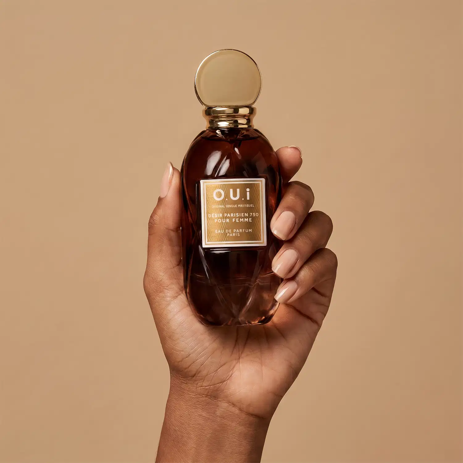 Frasco de item de perfumaria da marca O.U.i, com rótulo dourado, sendo segurado por uma mão feminina sobre fundo bege.
