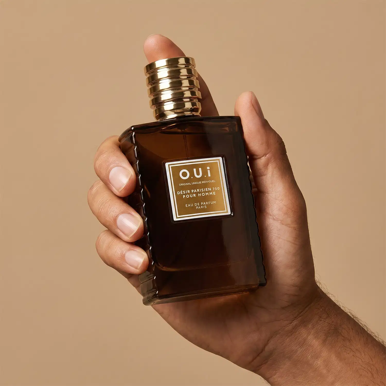 Recipiente de item de perfumaria da marca O.U.i em mãos masculinas sobre fundo bege, com detalhes dourados na embalagem.