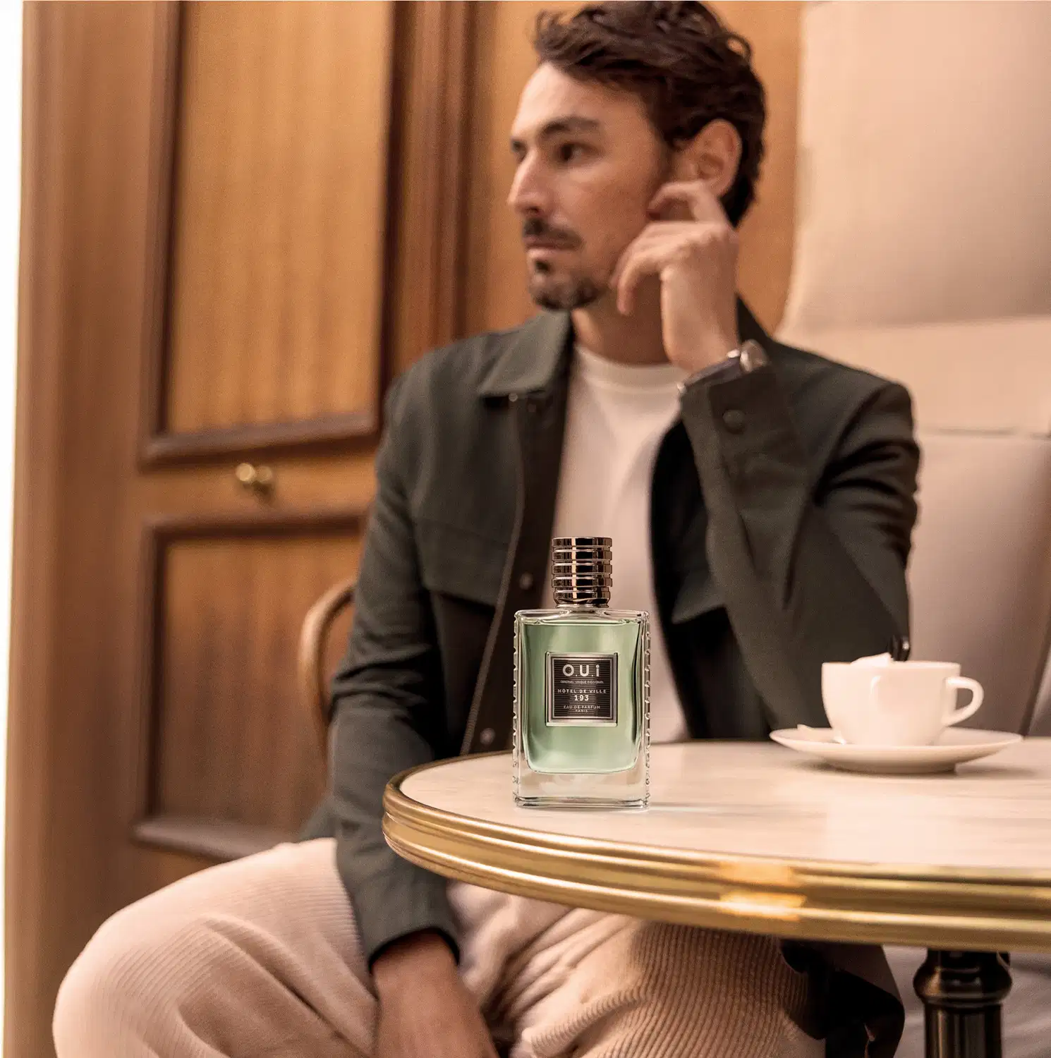Um homem de terno cinza está sentado em uma mesa de madeira, com uma xícara de café e um frasco de perfumaria da marca O.U.i em primeiro plano.