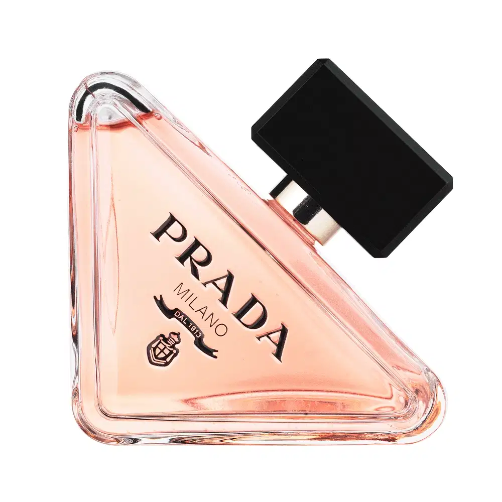 Frasco de perfume Prada com design triangular em vidro transparente, contendo líquido de cor rosa clara. O rótulo preto destaca o nome da marca "PRADA" em letras brancas.
