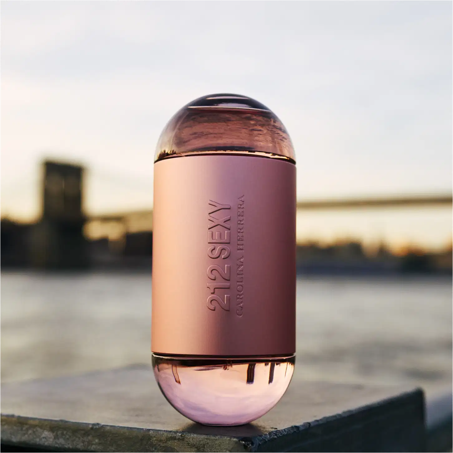Frasco de perfume Carolina Herrera 212 Sexy, com formato cilíndrico e tampa arredondada, em tons de rosa claro. O rótulo exibe o nome da fragrância em letras prateadas.