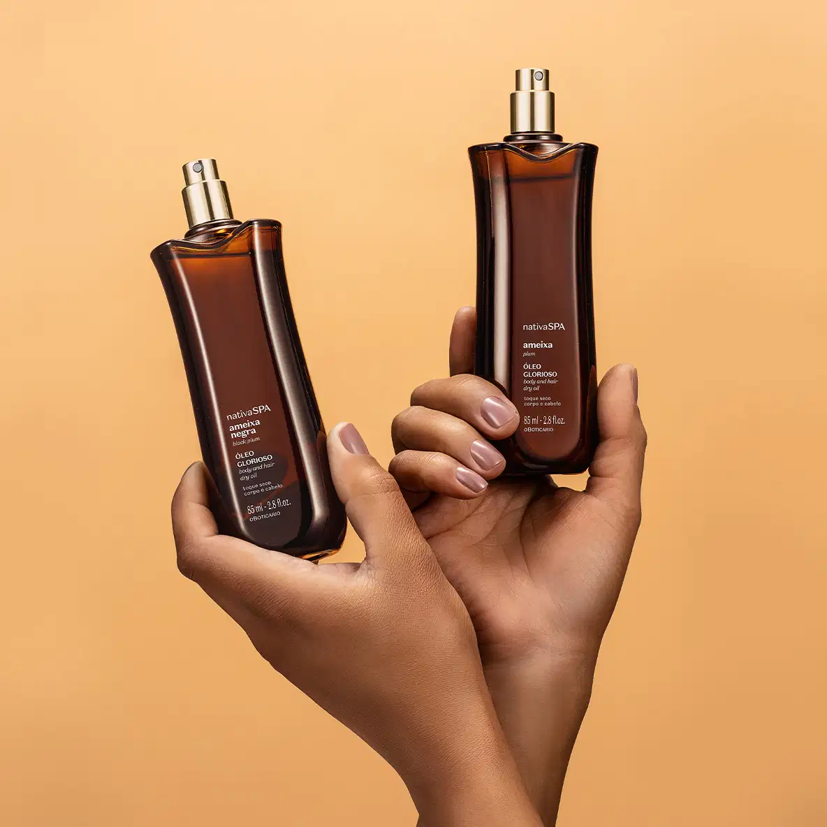 Mãos femininas segurando dois Frascos alongados na cor vinho escuro com borrifadores dourados e letras brancas de óleo multifuncional glorioso corpo e cabelos Nativa SPA Ameixa Plum em fundo laranja.