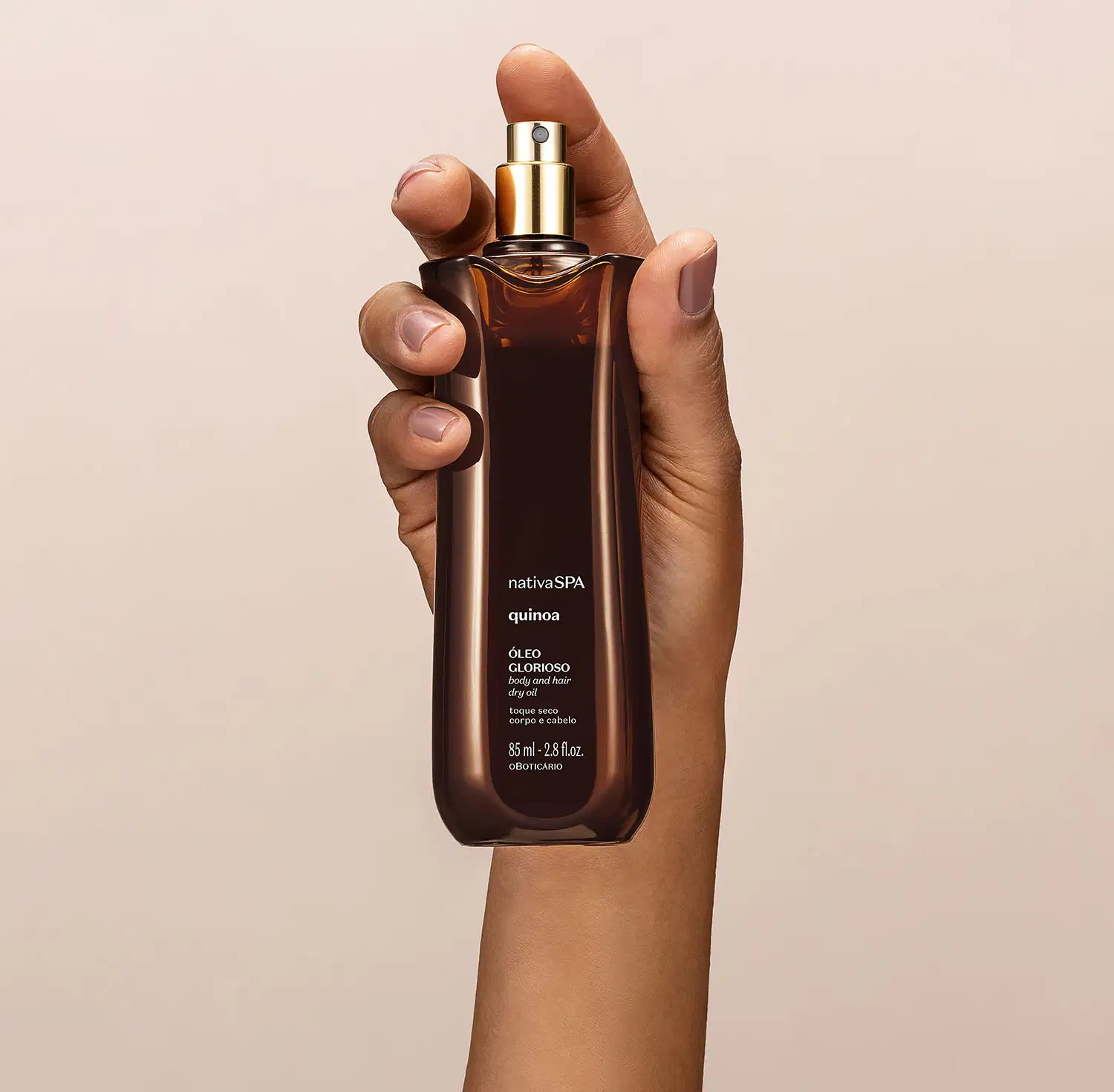mão segurando um vidro alongado marrom com pulverizador dourado e letras brancas contendo óleo multifuncional glorioso corpo e cabelos da marca Nativa SPA Quinoa em fundo bege.