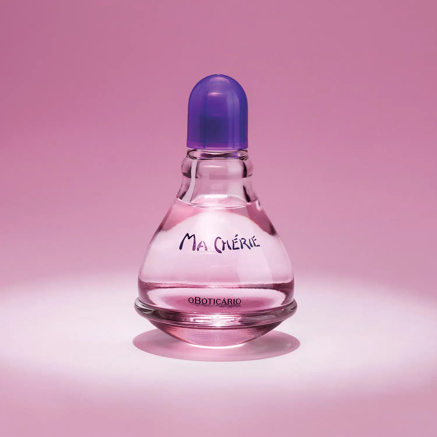Vidro transparente com fundo arredondado, líquido rosa, tampa e letras azuis, contendo 100ml de desodorante colônia Ma Chérie, posicionado em superfície rosa com sombra refletida, fundo rosa uniforme.