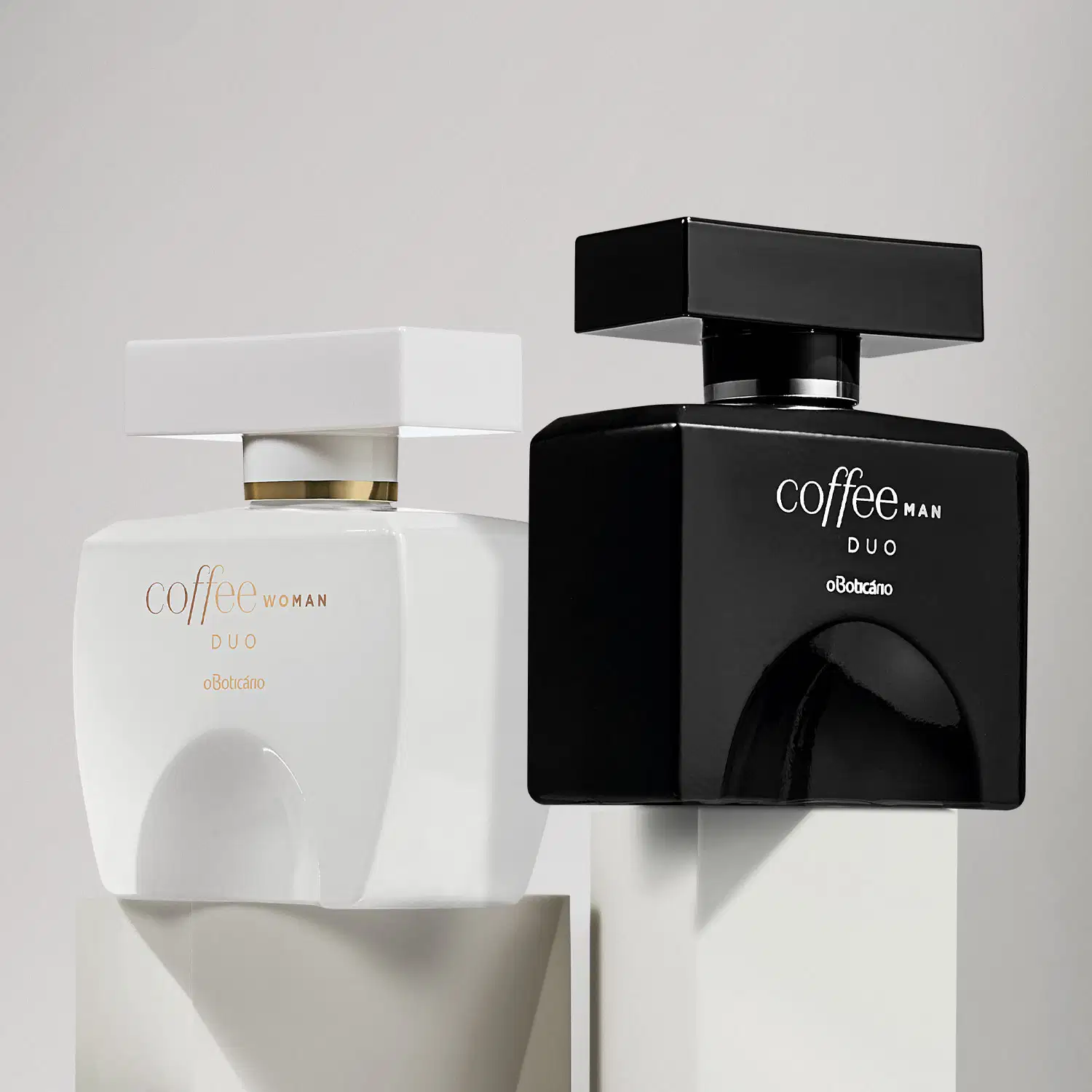 Dois vidros quadrados com tampa retangulares, detalhes e letras douradas, contendo desodorante colônia Coffee Duo, sendo um preto da linha Man e outro branco da linha Woman sobre plataformas brancas em fundo cinza.