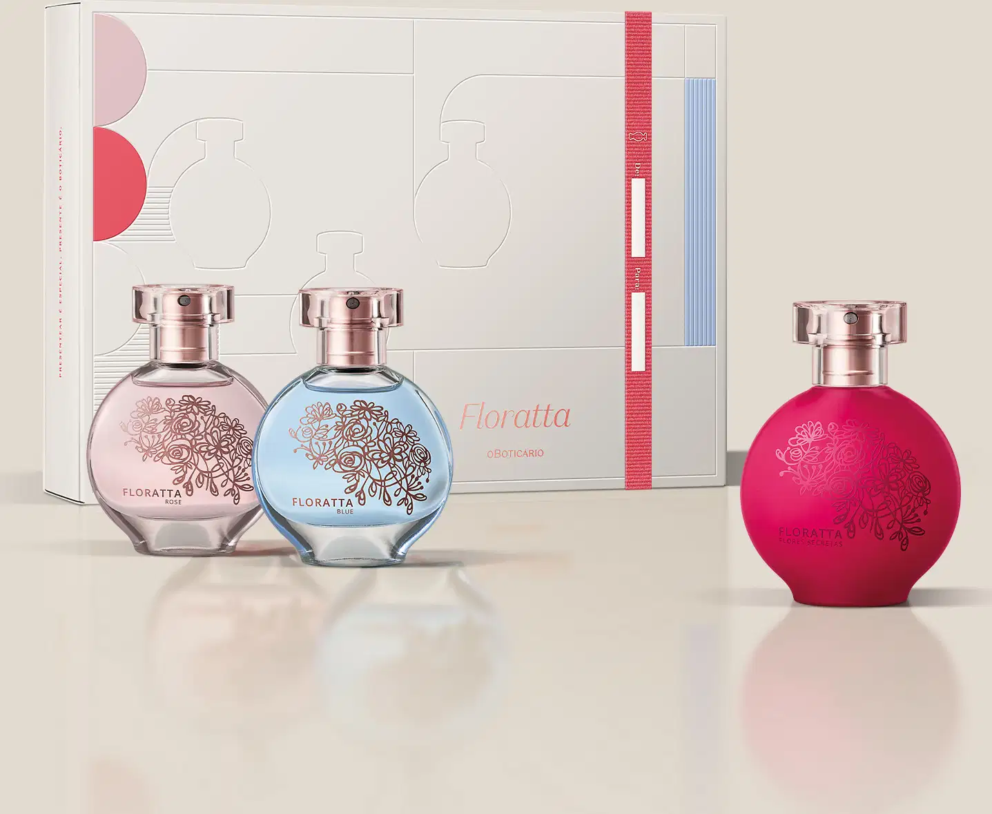 Três vidros redondos com florais em relevo, borrifador dourado e tampa cristal de desodorante colônia Floratta, sendo um rosa transparente da linha Rose, um azul transparente da linha Blue e um vermelho da linha Red, acompanhados de uma caixa de presente.
